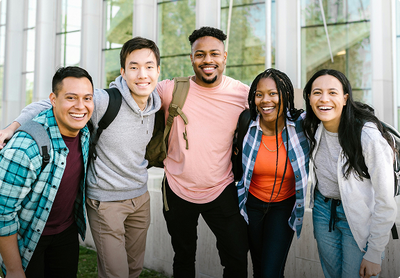 A diverse group of five friends standing together with backpacks, smiling outdoors.