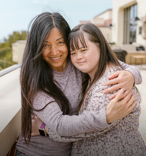 A woman embraces a young girl on a balcony, conveying warmth and affection in a serene outdoor setting.