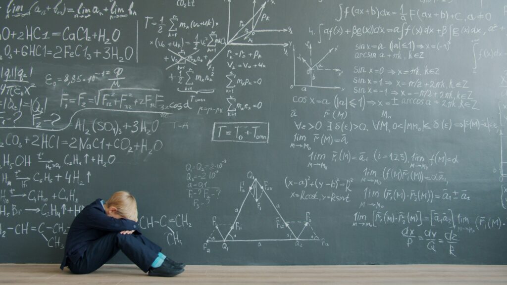 Child sitting on the floor in front of a classroom chalkboard, appearing overwhelmed by academic demands.